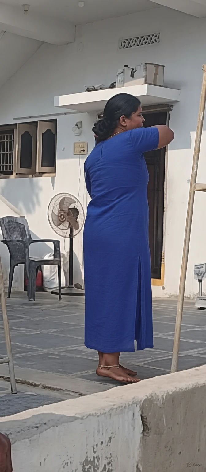 A woman in a vibrant blue dress stands barefoot on a tiled patio adjusting her hair near a white wall with a standing fan and plastic chair.