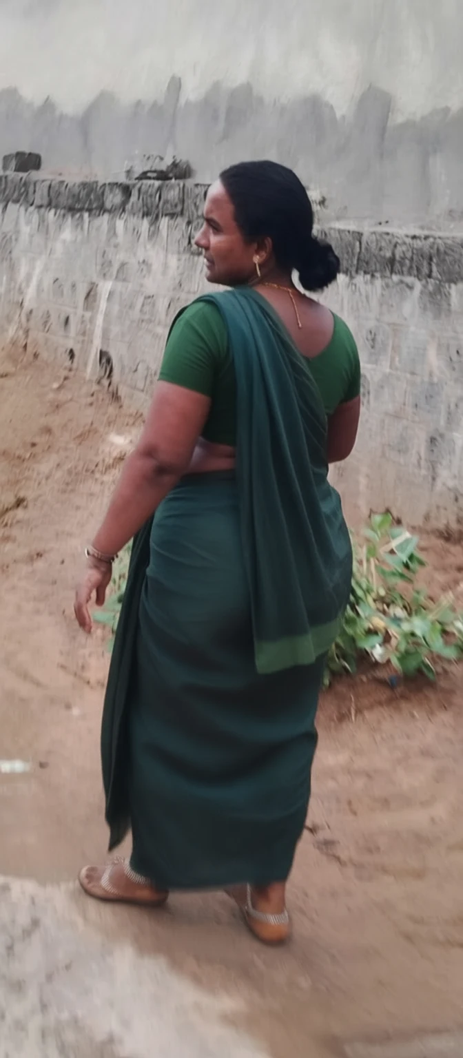 A woman in a dark green sari walks away on a dirt path beside a weathered concrete wall.