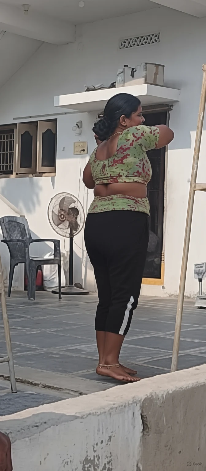 A woman in a green patterned top and black pants stands barefoot on a tiled patio adjusting her hair near a white wall with a standing fan and plastic chair.