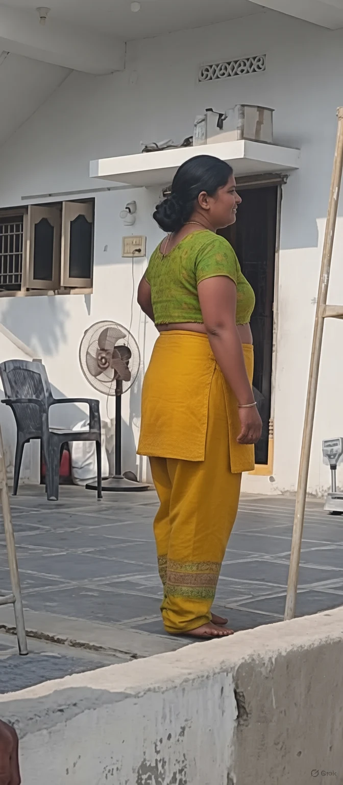 A woman in a green blouse and yellow pants stands barefoot on a tiled patio beside a standing fan and plastic chair.