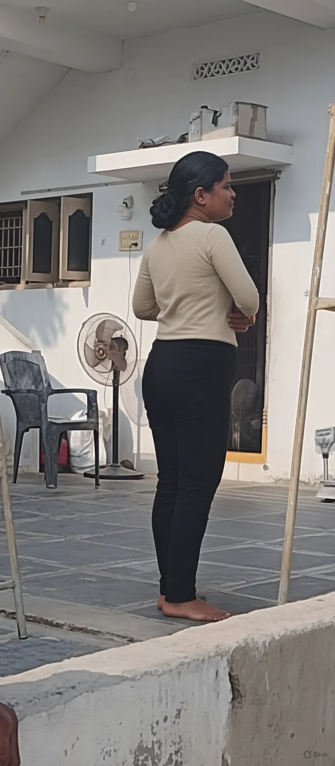 A woman in beige top and black pants stands barefoot on a tiled porch looking out with arms crossed near a standing fan and plastic chair.