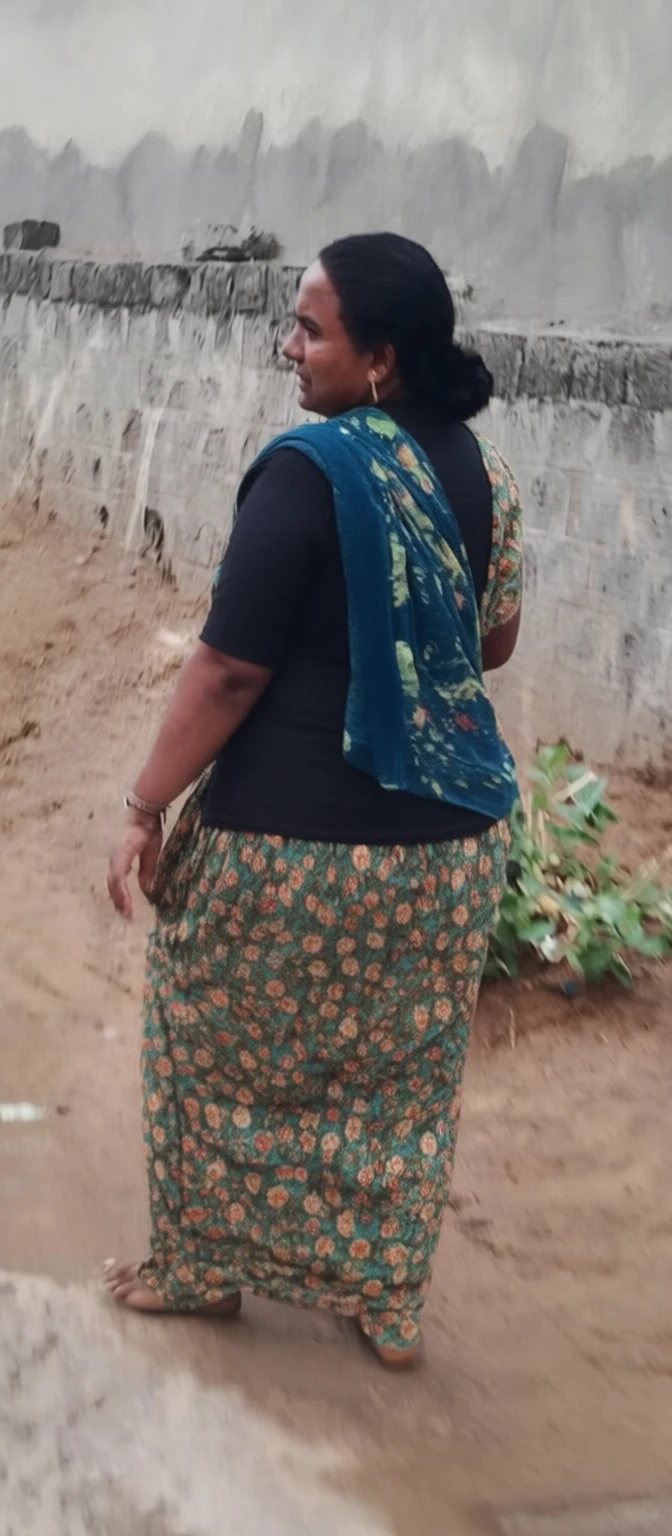A woman in a floral green skirt and blue patterned shawl walks barefoot on a dirt path beside a weathered concrete wall.