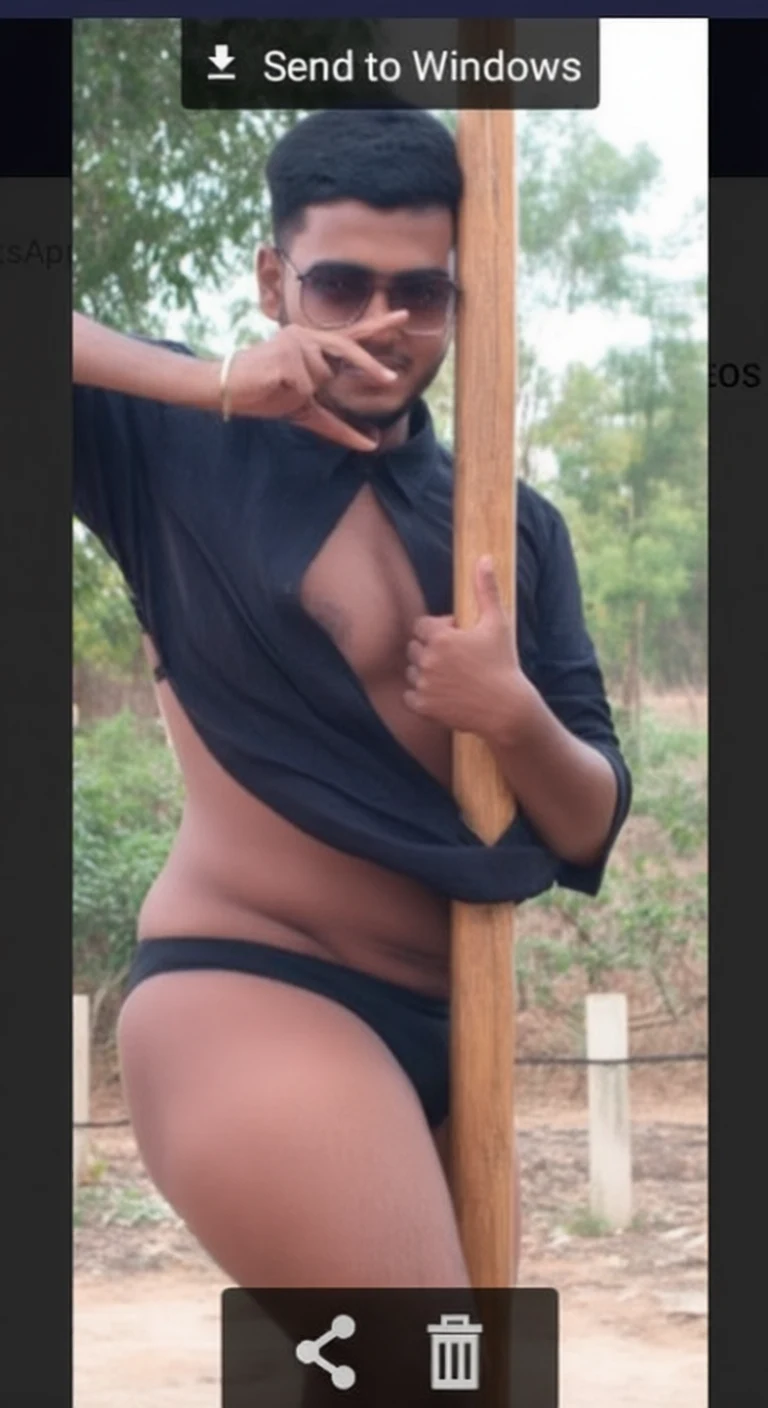 A man in black shirt and underwear leans against a wooden pole outdoors with trees in background.