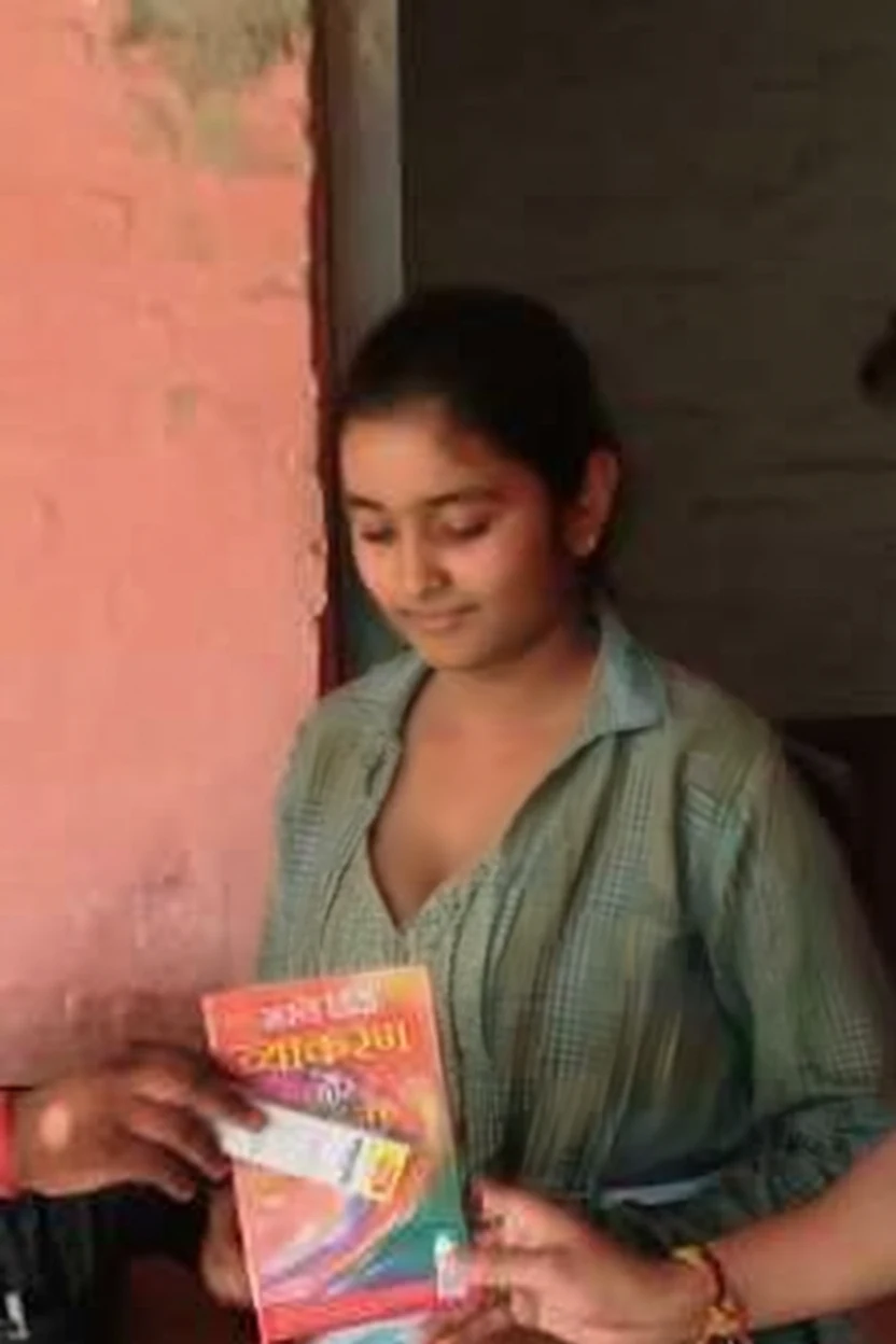 A young woman in a green checkered shirt smiles gently while holding a colorful book against a faded pink wall.